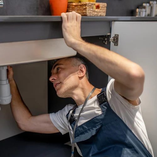 Encanador profissional com camiseta branca e macacão azul focado no trabalho, com o corpo parcialmente inserido embaixo do armário da pia para inspecionar e ajustar as conexões hidráulicas do sifão.