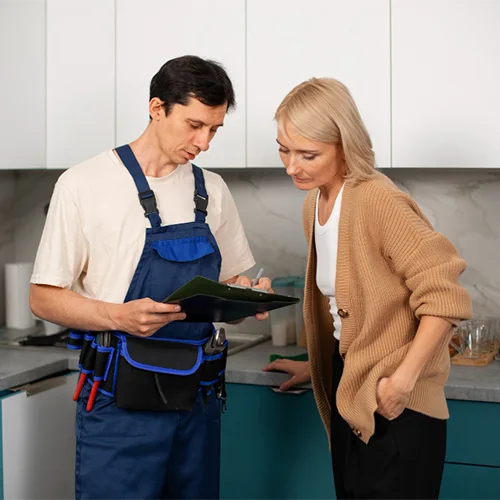 Profissional de serviços residenciais com uniforme azul e cinto de ferramentas conversando com uma cliente na cozinha enquanto avalia um orçamento em uma prancheta.