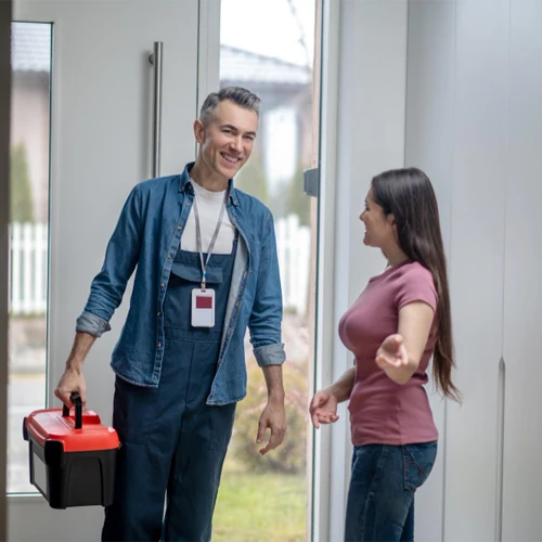 Profissional de manutenção residencial sorridente, vestindo macacão e crachá de identificação, chegando à porta de uma cliente e carregando uma caixa de ferramentas vermelha e preta.