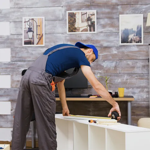 Montador de móveis profissional usando macacão cinza e boné azul, inclinado sobre um móvel de MDF branco em montagem para tirar medidas precisas com uma trena manual.
