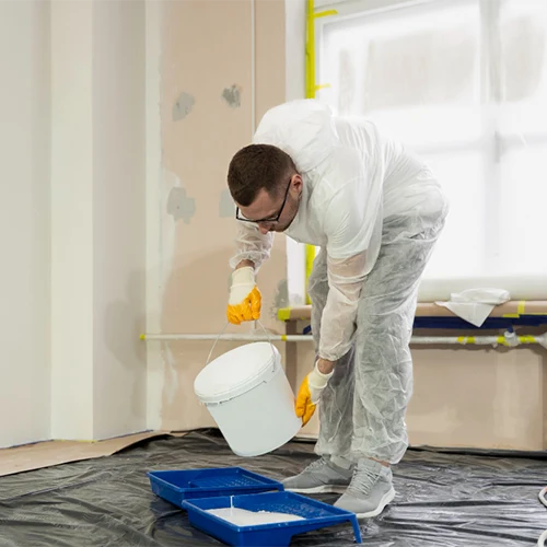 Pintor residencial vestindo macacão de proteção branco, óculos de segurança e luvas amarelas, inclinado para despejar tinta branca de um balde em uma bandeja de pintura azul sobre o piso protegido com lona preta.
