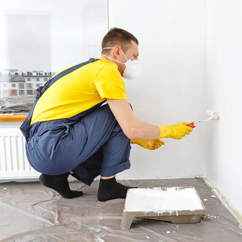 Pintor residencial de perfil, usando camiseta amarela, macacão azul, luvas e máscara de proteção, agachado sobre o piso protegido com lona enquanto aplica tinta branca na parede utilizando um rolo de pintura.